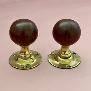 May include: A pair of door knobs featuring spherical dark brown wooden heads attached to polished brass circular bases. Each base includes three pre-drilled screw holes for mounting, showcasing a metallic gold finish and a smooth wood grain texture.