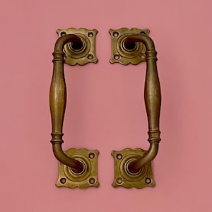 Puede incluir: Un par de tiradores de puerta de latón antiguo con detalles ornamentados. Los tiradores están fijados a una pared rosa.
