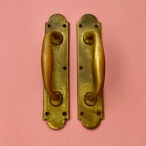 May include: Two antique brass door handles with a curved design. The handles have a polished finish and are mounted on a flat surface.