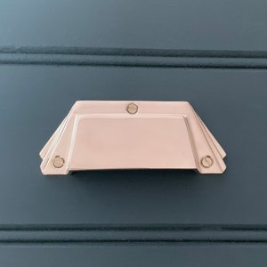 Può includere: Una maniglia per armadio in oro rosa con forma rettangolare e una parte superiore e inferiore leggermente arrotondata. La maniglia ha una finitura liscia e lucida e presenta due piccole viti rotonde su ciascun lato.