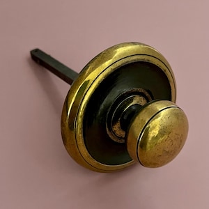 May include: A vintage brass bottle stopper with a dark wooden stem. The stopper features a circular design with a rounded knob on the end. The brass has a warm, aged patina, suggesting its antique nature.