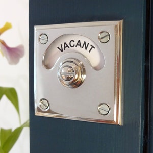 Puede incluir: Una perilla de puerta cromada con un letrero blanco redondeado que dice "VACANT". La perilla está montada en una puerta azul oscuro.