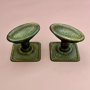 May include: A pair of matching door knobs with an oval-shaped handle and a square base plate. Both pieces feature a dark, aged brass finish with a textured, hammered surface pattern on the handle and the plate, including four screw holes.