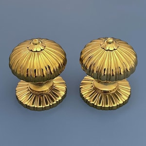 Pode incluir: Dois puxadores dourados para armários. Cada puxador apresenta um topo arredondado e canelado e uma base detalhada. Os puxadores são feitos de um material metálico brilhante e são colocados sobre um fundo azul liso.