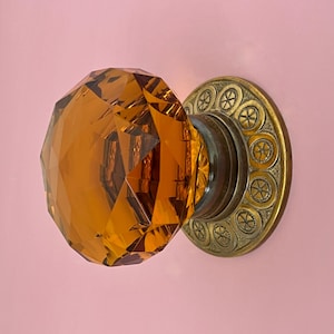 May include: Amber-colored, faceted glass doorknob with a brass backplate. The doorknob has a round, jewel-like appearance. The backplate is circular with a repeating pattern of small, stylized floral designs. The doorknob is mounted on a pink wall.