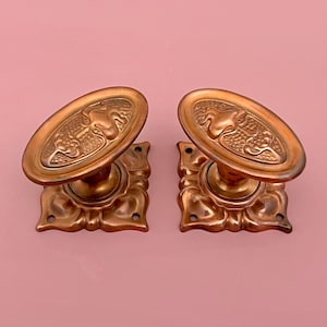 May include: Two antique bronze door knobs. Each knob features an oval handle with an embossed design and a decorative square base. The knobs are a warm copper color and are set against a pink background.