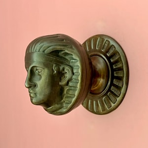May include: A dark bronze-toned metal door knob featuring a sculpted relief of a human head in profile. The knob is attached to a circular backplate with a decorative radial ribbed pattern around the outer edge, creating a textured appearance.