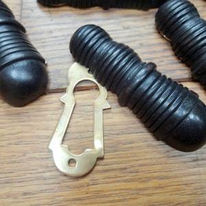 Può includere: Un set di cinque pomelli per porte in legno nero con una placca per buco della serratura in ottone. I pomelli hanno un design a coste.