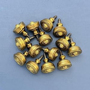 May include: A group of 12 gold-toned metal cabinet knobs with a decorative design. Each knob has a screw for attaching to a cabinet door.