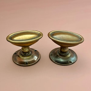 Peut inclure: Une paire de poignées de porte ovales en laiton avec une finition métallique lisse et une tige centrale reliée à une plaque de base circulaire avec des trous de vis visibles. Le laiton présente un ton brun doré chaud avec des nuances.