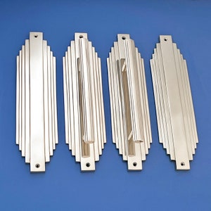 Puede incluir: Cuatro tiradores de puerta plateados con un diseño Art Deco. Cada tirador tiene una forma rectangular con una ranura vertical en el centro.