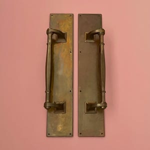 Puede incluir: Dos tiradores de puerta antiguos de bronce. Cada uno tiene una placa trasera rectangular, un mango largo y curvado y placas de montaje cuadradas. El bronce tiene un aspecto envejecido, con variaciones de color y textura.