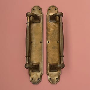 Puede incluir: Dos manijas de puerta antiguas de latón, con una placa trasera rectangular y un asa curva. El latón tiene un aspecto envejecido y deslustrado. El fondo es de color rosa liso y apagado.