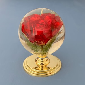 May include: A red rose encased in a clear, spherical glass. The rose is set on a gold-colored, ornate stand. The base of the stand is round, and the stem is detailed. The background is a solid, muted blue.