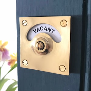 May include: A brass door knob with a round, rotating indicator that says "VACANT" in black lettering. The knob is mounted on a dark blue door.