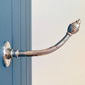 May include: A silver metal hook with a decorative finial. The hook is mounted on a wall and is curved.
