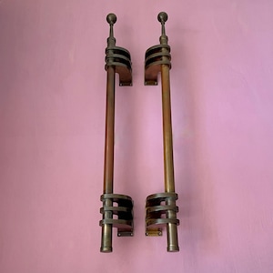 May include: Pair of vintage bronze door handles. Each handle features a long, cylindrical shaft with decorative bronze elements, including a ball finial at the top and a series of circular bands. The handles are mounted on rectangular plates.