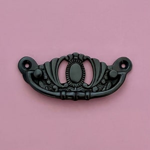 Può includere: Tiramaniglia per cassetto in metallo nero con un design ornamentale. La maniglia ha una forma ovale arrotondata al centro ed è circondata da un motivo floreale decorativo.