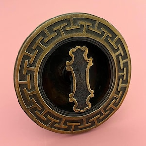 May include: A bronze-toned, circular cabinet pull with a geometric border design. The center features a dark, ornate handle. The pull is set against a soft pink background.