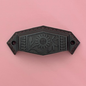 Puede incluir: Un tirador de cajón de metal marrón oscuro con un diseño de girasol y hojas. El herraje decorativo tiene forma rectangular con un patrón floral en el centro y un borde decorativo. El tirador está sobre un fondo rosa.