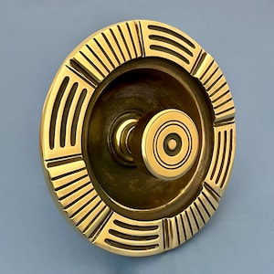 May include: A brass door knob with a circular design. The knob features a central round handle with concentric circles and is surrounded by a decorative ring with vertical and curved lines. The metal has a polished, golden finish.