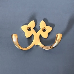 Puede incluir: Un gancho doble dorado con un diseño floral. Los ganchos son curvos y tienen una forma de flor decorativa en la parte superior.