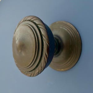 May include: A close-up shot of a bronze door knob. The knob has a circular design with a detailed, textured edge. The background is a solid light blue. The door knob is a classic piece of hardware.