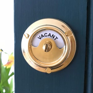 May include: A brass door knob with a white sign that says "VACANT" on it. The knob is mounted on a dark blue door.