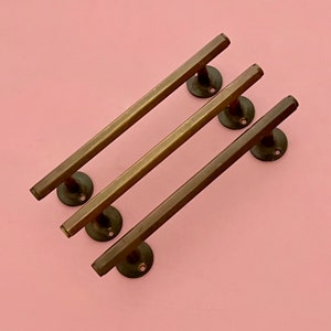 Può includere: Tre maniglie per armadi in ottone con forma rettangolare e un bordo leggermente arrotondato. Le maniglie sono disposte in modo sfalsato su uno sfondo rosa.