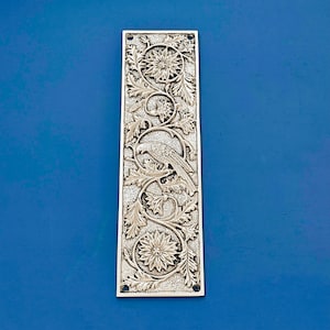 Può includere: Una placca per porta in argento con un designo ornamentale che presenta un uccello posato su un ramo con foglie e fiori.