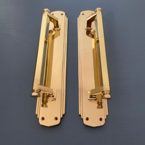 May include: Two polished brass door handles with a rectangular shape and a long, straight lever. The handles have a decorative Art Deco design with geometric lines and a smooth, shiny finish.