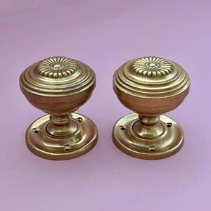May include: A pair of round brass door knobs featuring a central floral motif with concentric ridged detailing. Each knob is attached to a circular mounting plate with three visible screw holes, showcasing a polished metallic finish.