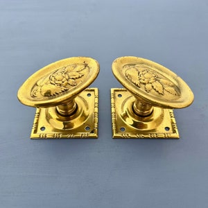 May include: A pair of antique brass door handles with an oval-shaped design featuring a floral motif. The handles are mounted on square backplates with a decorative border.