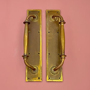 Puede incluir: Un par de tiradores de puerta de latón antiguo con diseños ornamentados. Los tiradores están unidos a placas rectangulares con un borde decorativo.