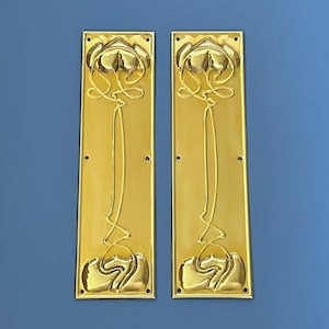 Puede incluir: Dos placas de puerta rectangulares doradas con un diseño floral Art Nouveau. Cada placa presenta una flor estilizada en la parte superior e inferior, conectada por un tallo fluido. Las placas están montadas sobre un fondo azul.