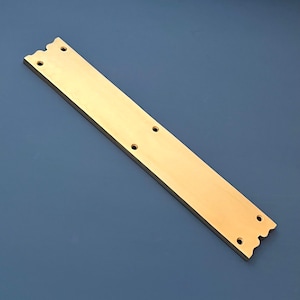 Puede incluir: Una placa de metal larga, plana y de color dorado con bordes redondeados y varios agujeros. La placa tiene una superficie ligeramente texturizada.