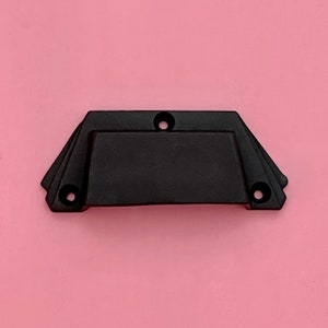 Può includere: Tiramaniglia per cassetto in metallo nero con forma rettangolare e bordi arrotondati. La maniglia ha una sezione centrale leggermente rialzata e quattro piccoli fori.