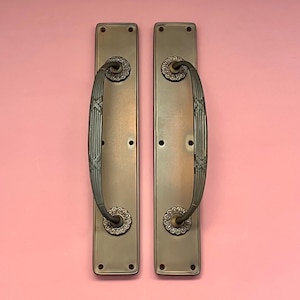 Puede incluir: Un par de manillas de puerta de estilo antiguo con detalles ornamentados. Las manillas son de metal oscuro con acabado cepillado. Tienen una placa posterior rectangular y una manija de palanca curva.