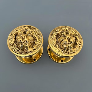 以下が含まれることがあります： ライオンの頭のデザインが施された金色のドアノブのペア。ライオンの頭は毛皮と顔の特徴が詳細に描かれています。
