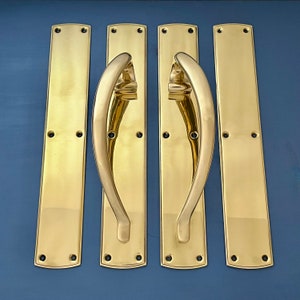 May include: Set of four polished brass door pulls and plates. Two curved handle pulls are paired with two rectangular plates, all with mounting holes. The brass has a bright, reflective finish, suitable for interior or exterior doors.