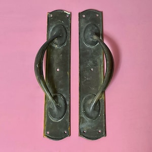 May include: A pair of antique door handles with a curved design. The handles are made of brass and have a dark patina. They are mounted on rectangular plates with decorative edges.