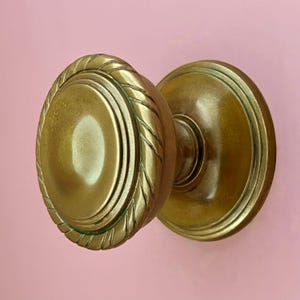 May include: A pair of antique brass door knobs. The knobs have a warm, golden hue and a classic design. One knob features a rope-like border around the oval face. The other knob is a simple, round design. The background is a soft pink.