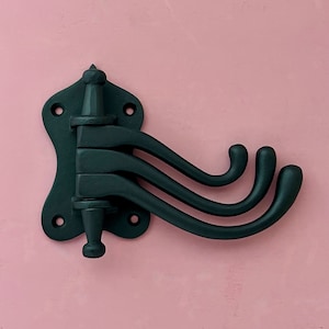 Puede incluir: Perchero de pared de metal negro con tres ganchos. El perchero tiene forma de S estilizada con un remate decorativo en la parte superior.