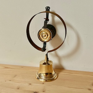 May include: A brass bell with a round, black metal frame and a brass striker. The bell is mounted on a wooden surface.