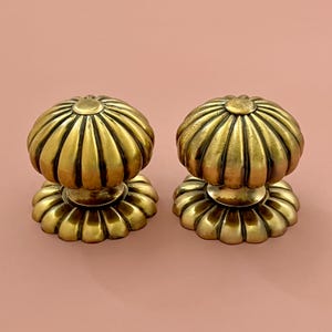 Peut inclure: Deux boutons de meuble anciens en laiton avec un design cannelé en forme de citrouille. Les boutons ont une teinte dorée chaude et sont posés sur un fond rose clair. La base de chaque bouton a un bord festonné.