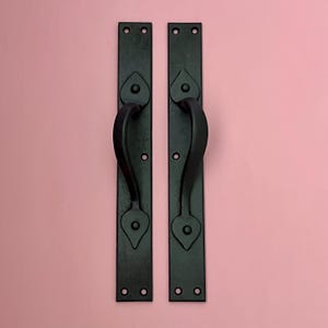 Könnte beinhalten: Zwei schwarze Metall-Türgriffe mit herzförmigem Design. Jeder Griff hat eine lange, rechteckige Rückplatte mit einem gebogenen Griff. Die Griffe sind auf einem rosa Hintergrund montiert.