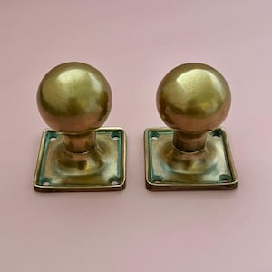 May include: Two matching brass door knobs with spherical heads and square base plates. Each plate features four circular screw holes at the corners. The metal has a polished finish with visible signs of aging and patina in the recessed areas.