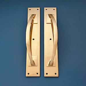 Puede incluir: Dos tiradores de puerta dorados con un diseño curvo. Los tiradores son rectangulares y tienen un acabado pulido.