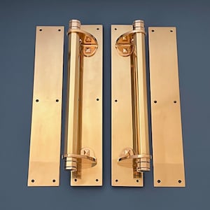 Puede incluir: Un par de manillas de puerta de latón pulido con una placa posterior rectangular y una palanca larga y cilíndrica. Las manillas tienen un diseño geométrico y están montadas en una pared.