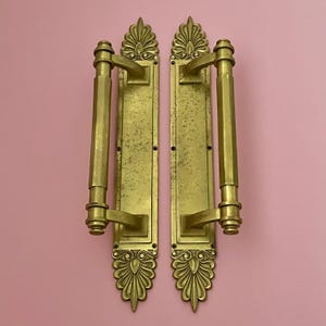 Puede incluir: Dos manijas de puerta antiguas de latón con diseños florales y geométricos ornamentados. Las manijas son rectangulares con una barra vertical y una parte superior e inferior decorativas. Fondo rosa liso.